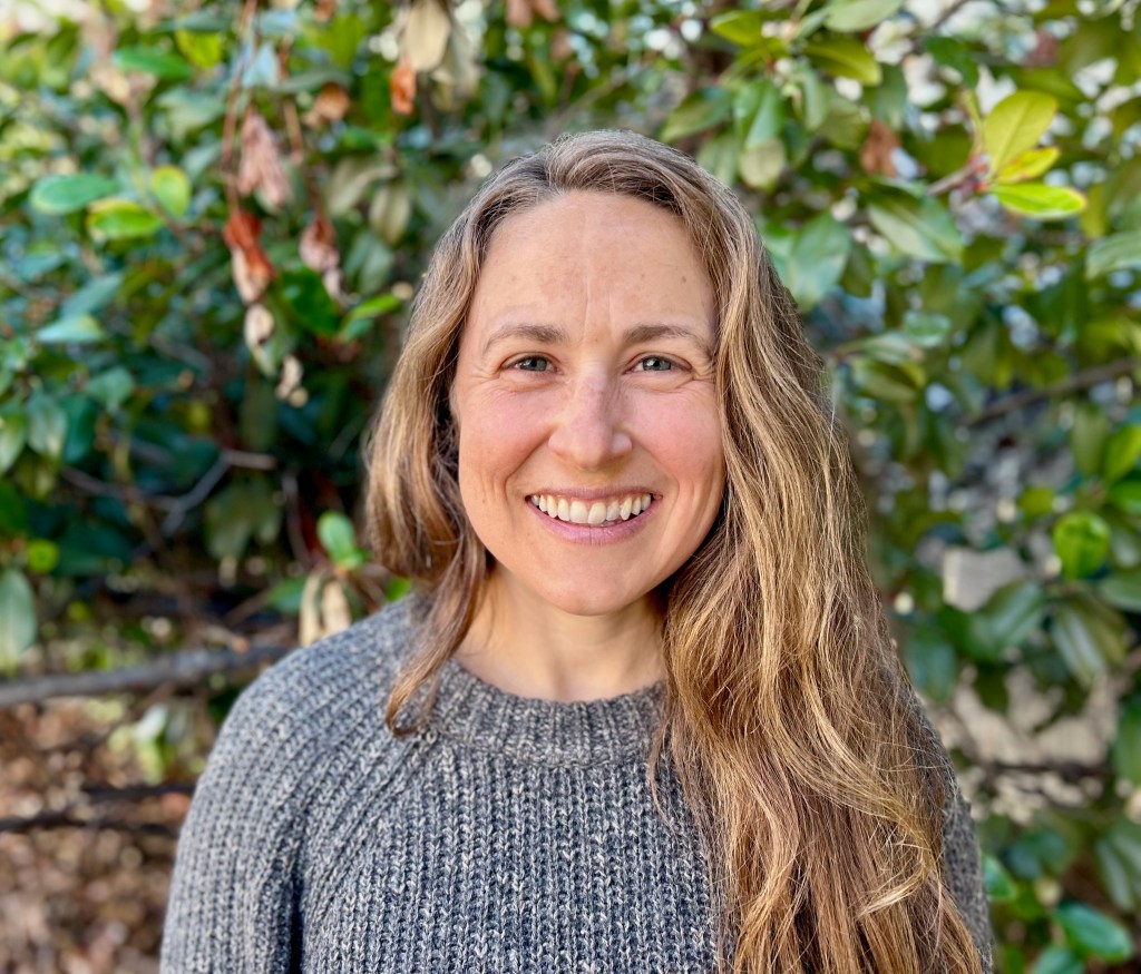 Smiling woman with long hair wearing a gray sweater, standing outdoors among green foliage.